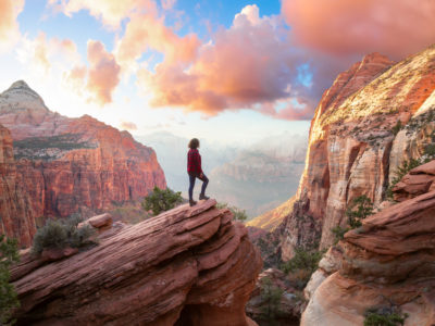 Adventurous Woman at the edge of a cliff is looking at a beautiful landscape view in the Canyon during a vibrant sunset. Taken in Zion National Park, Utah, United States. Sky Composite.
